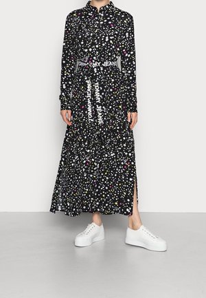 Femme portant une longue robe noire à motif floral coloré, ceinturée avec un texte "Tommy Jeans", assortie à des baskets blanches à plateforme.