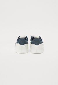 Hvite sneakers med en glatt læroverdel og marineblå detaljer ved hælen. Har et rundt logo og en teksturert såle.
