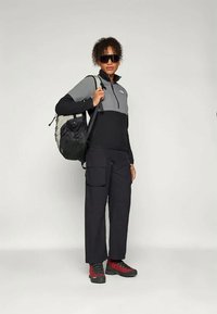 Mulher vestindo uma jaqueta com fecho éclair preta e cinza, calças cargo pretas, botas de caminhada vermelhas, óculos de sol escuros e carregando uma mochila preta e cinza.