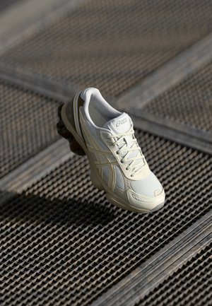 Weißer und beiger Asics-Laufschuh mit Mesh- und Lederdetails, der über einem strukturierten Metallboden schwebt.