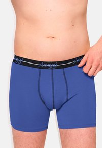 Blaue Baumwollboxershorts mit einem schwarzen elastischen Bund, der ein Logo trägt. Der Stoff ist glatt mit kontrastierenden Nähten.