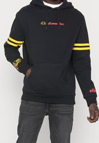 Felpa nera con strisce gialle doppie sulle maniche, testo "Cobra Kai" in rosso sul petto, testo giallo e rosso sugli avambracci, indossata con jeans scuri.
