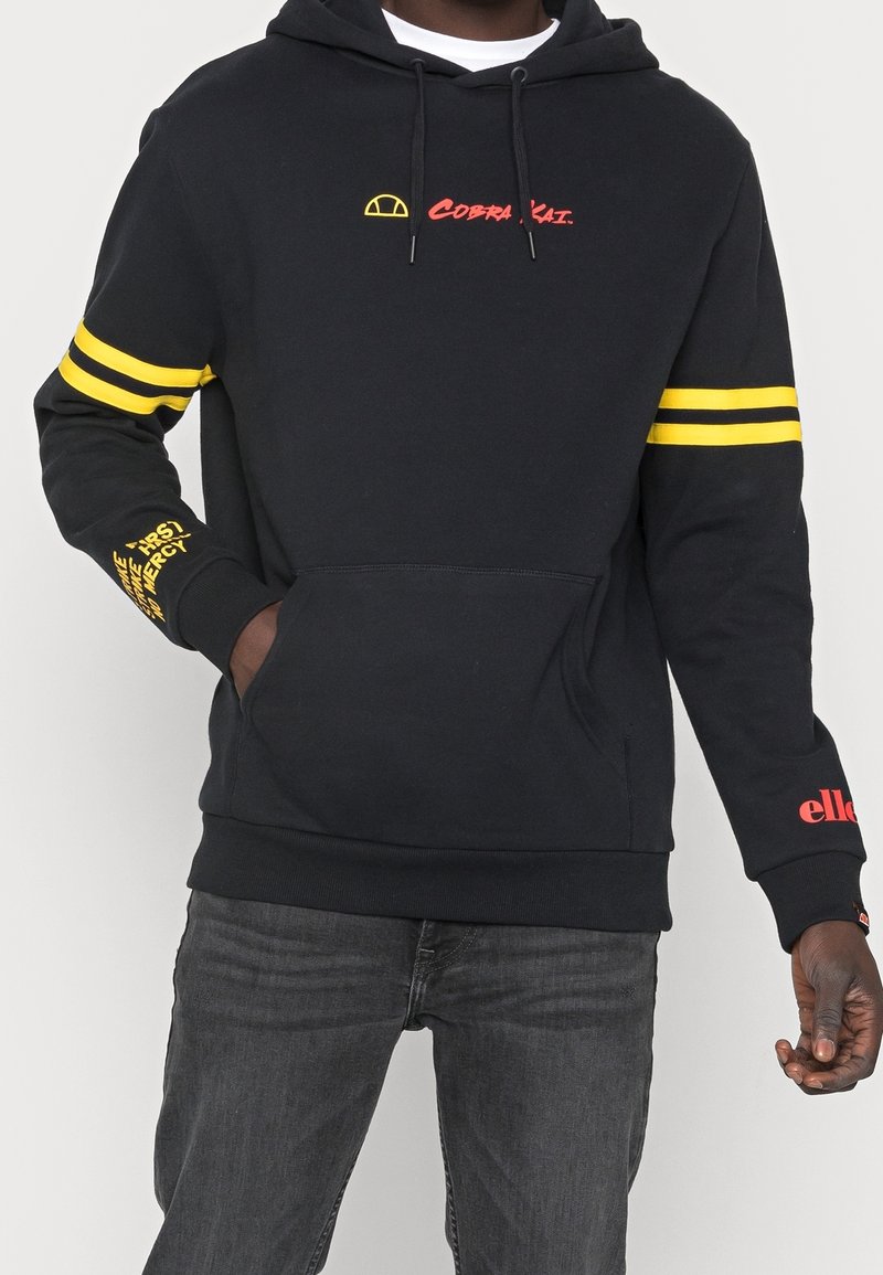 Felpa nera con strisce gialle doppie sulle maniche, testo "Cobra Kai" in rosso sul petto, testo giallo e rosso sugli avambracci, indossata con jeans scuri.