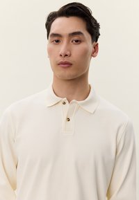 Νέος ενήλικας με κοντά σκουρόχρωμα μαλλιά, ντυμένος με κρεμ μακρυμάνικο πόλο πουκάμισο, σε απλό φωτεινό φόντο.