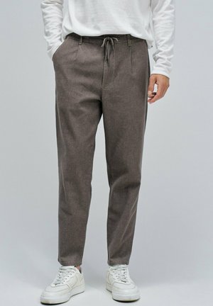 Hombre con pantalones marrones con cordón, camisa blanca de manga larga y zapatillas blancas, de pie con una mano en el bolsillo.