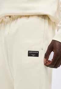 Κοντινή λήψη ενός ατόμου που φοράει κρεμ παντελόνι φόρμας με μαύρη ετικέτα Björn Borg και ελαστική μέση με κορδόνι.