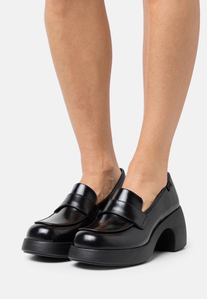 Mocassins en cuir noir brillant avec des talons hauts et épais, portés pieds nus, présentés de face et de profil sur un fond clair.