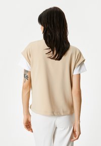 Top beige senza maniche con una finitura texturizzata, vestibilità morbida e una maglietta bianca visibile alle maniche. Cuciture semplici lungo l'orlo.