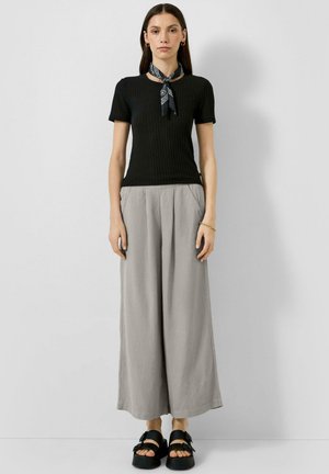 Frau trägt ein schwarzes kurzärmeliges Oberteil, graue Wide-Leg-Hosen, schwarze Sandalen und ein schwarzes gemustertes Halstuch, steht vor einer schlichten Wand.