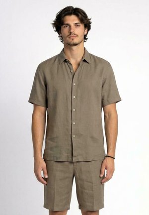 Giovane uomo che indossa una camicia marrone chiaro a maniche corte abbottonata e pantaloncini fino al ginocchio abbinati, in piedi contro uno sfondo bianco semplice.