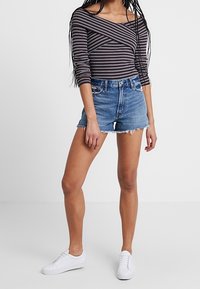 Top de hombros descubiertos a rayas con mangas largas, combinado con pantalones cortos de mezclilla azul de cintura alta con dobladillo deshilachado, y zapatillas deportivas blancas.