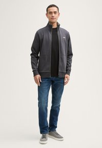 Chándal gris con cremallera y cuello alto, combinado con una camiseta negra, jeans azules y zapatillas grises. Presenta dos bolsillos frontales y una textura suave.