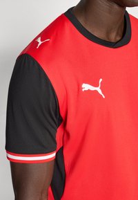 Piros sportpóló fekete ujjakkal, fehér Puma logóval a mellkason és csíkos díszítéssel a mandzsettákon. Légáteresztő anyagból készült.