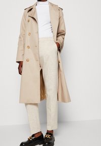 Béžový trenčkot s dvouřadým zapínáním, pod ním světlejší svetr, krémové kalhoty a černé mokasíny se zlatým řetízkovým detailem.