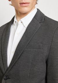 Czarno-biała teksturowana marynarka z klapą z wycięciem, zapinana na jeden guzik, z kieszonką na piersi, noszona na białej koszuli z kołnierzykiem.