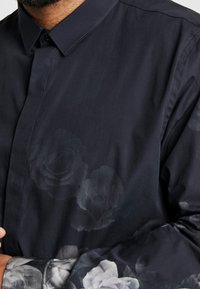 Chemise bleu marine foncé avec un motif floral subtil sur la poitrine et la manche, portée par une personne avec une barbe visible et une main près du col.