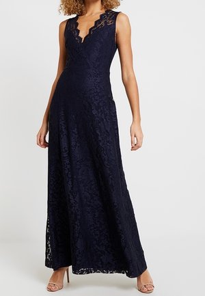 Femme portant une robe longue en dentelle bleu marine sans manches avec un encolure en V festonnée et des sandales beige à talons hauts.