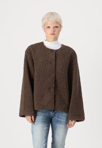 Veste marron, duveteuse, à boutons avec un col rond et des manches larges, portée sur un haut blanc à col montant, assortie avec un jean bleu.