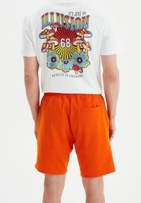 T-shirt blanc avec un motif graphique coloré représentant des champignons et des fleurs. Short orange avec une poche arrière, en tissu lisse.