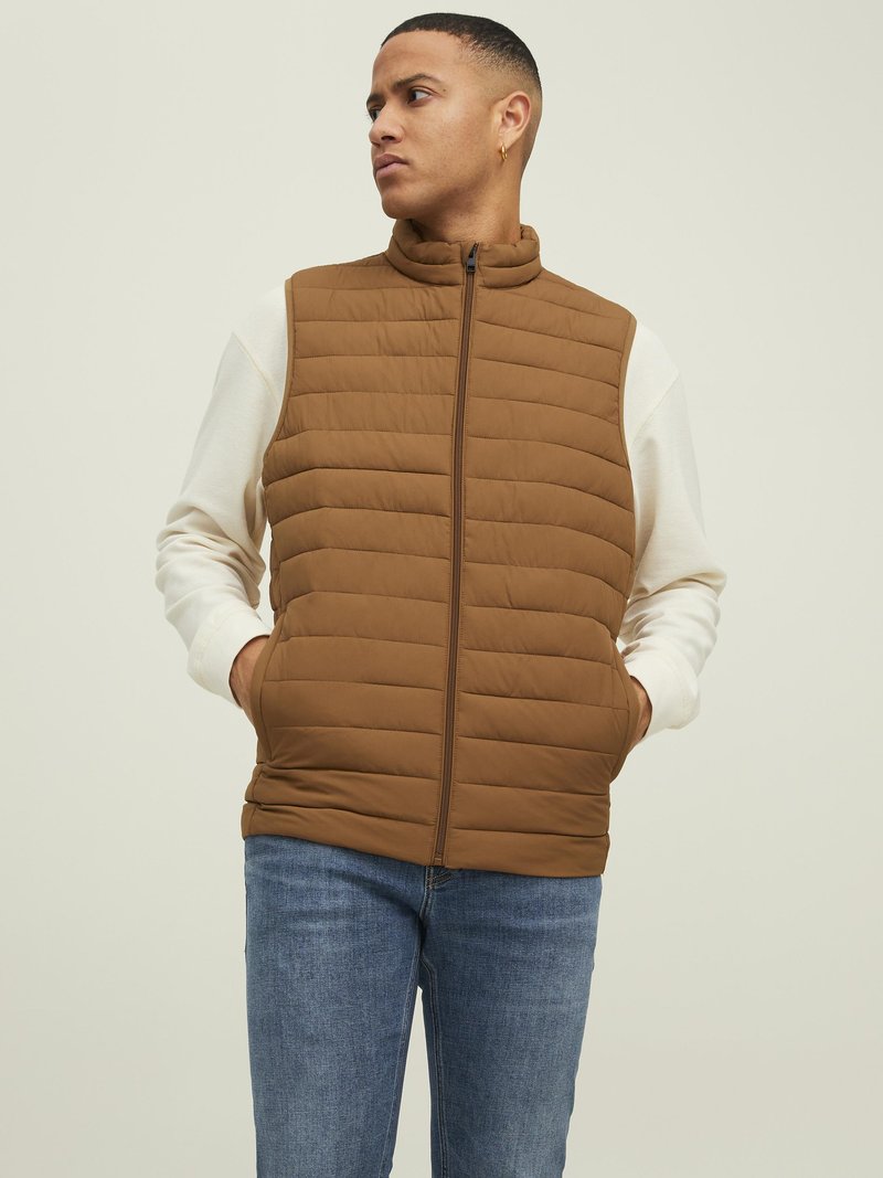 Bruine gewatteerde vest met hoge kraag, rits aan de voorkant en zijzakken, gedragen over een crème lange mouw shirt, gecombineerd met blauwe spijkerbroek.