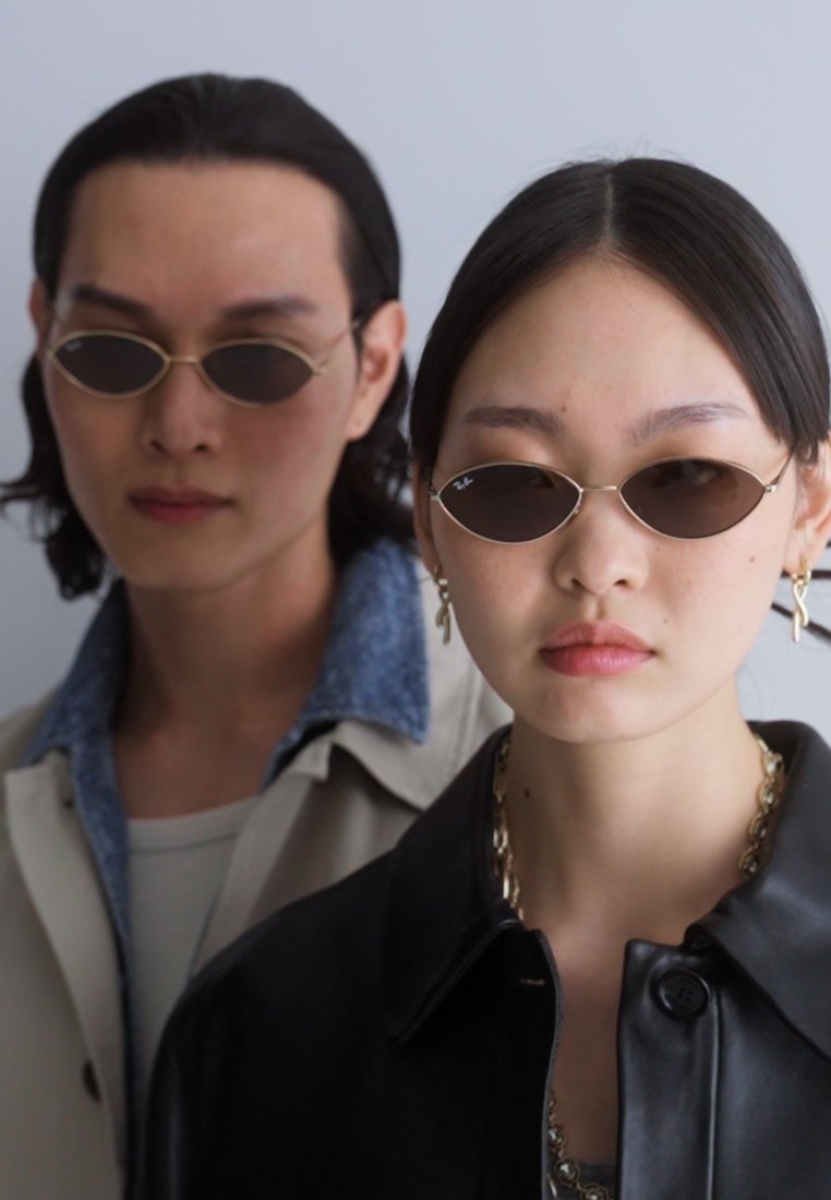 Lunettes de soleil à monture ovale fine de couleur sombre, portées par deux modèles. L'un porte une veste en cuir ; l'autre porte une couche extérieure avec un col en denim.