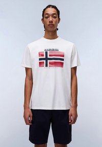 Maglietta bianca di cotone con grafica della bandiera norvegese in rosso, bianco e blu, con la scritta "NAPAPIJRI" sopra. Il modello indossa pantaloncini scuri.