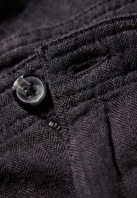 Svart herringbone-mönstrad tyg med en glänsande svart knapp och metallblixtlåsdetalj, som visar upp en texturerad väv och sömmar.