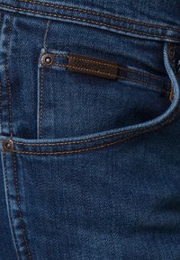 Vaqueros de mezclilla azul oscuro con un acabado texturizado, costuras doradas, una pequeña etiqueta de cuero y un bolsillo delantero con detalle de remache de metal.