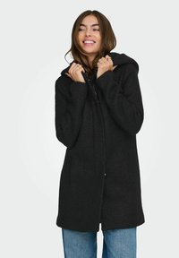 Manteau noir en mélange de laine avec capuche, fermeture éclair sur le devant et deux poches latérales ; présente une texture douce et une longueur mi-thigh.