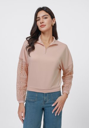 Pullover rosa claro com fecho de zip até ao quarto, com mangas em padrão de renda e uma textura suave, combinado com jeans azuis de cintura alta.