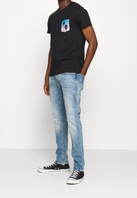 Camiseta negra de algodón con un bolsillo gráfico que muestra palmeras; combinada con vaqueros azul claro y zapatillas de lona negras.