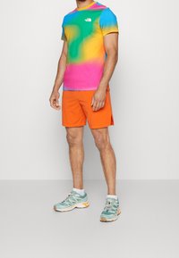 T-Shirt mit mehrfarbigem Farbverlauf in Blau-, Grün-, Gelb- und Pinktönen; orangefarbene Shorts; leichte türkisfarbene Laufschuhe mit weißen Akzenten.