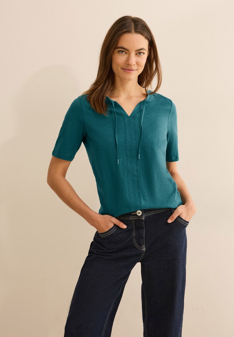 Top corto de color azul verdoso con cuello en V y cordones, combinado con pantalones de mezclilla oscuros que presentan costuras contrastantes y bolsillos laterales.