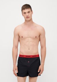 Schwarze Boxershorts mit einem roten HUGO-Bund, ausgestattet mit einem Knopfverschluss und Steppnähten. Glatte Textur mit maßgeschneiderter Passform.