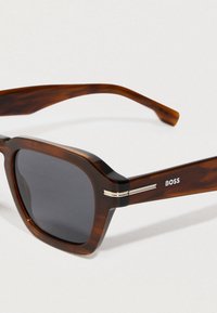 Καφέ καπιτονέ ορθογώνια γυαλιά ηλίου με σκούρους φακούς και ασημένιες τονισμένες γραμμές στους βραχίονες, με την ετικέτα "BOSS" στο πλάι.