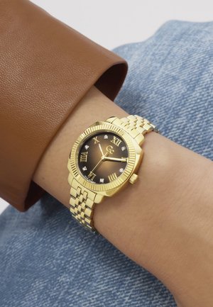 Reloj de pulsera de oro con esfera de degradado marrón, números romanos y marcadores de diamantes en la muñeca, usado con manga marrón y fondo de vaqueros azules.