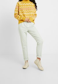 Osoba ubrana w żółto-różowy sweter we wzory, jasnobeżowe spodnie i kremowe wysokie trampki stojąca na białym tle.