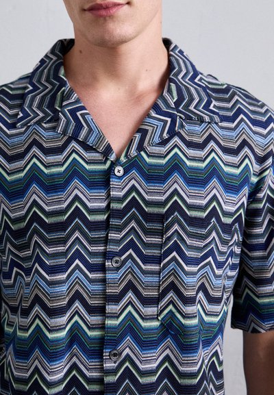 Uomo che indossa una camicia a maniche corte con bottoni, caratterizzata da un motivo a zigzag blu, verde e bianco, e colletto.