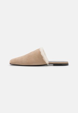Mule beige en daim à enfiler avec doublure en shearling blanc et semelle plate noire, vue de profil sur fond blanc.