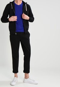 Person klädd i en blå t-shirt, svart zip-up hoodie, rullade svarta byxor och vita sneakers, stående mot en enkel ljus bakgrund.