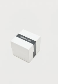 Boîte en carton blanc avec un bandeau en papier noir portant le texte "Calvin Klein" en blanc. La boîte a un design simple et une forme carrée.