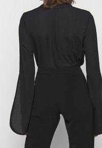 Blusa nera con maniche lunghe a campana trasparenti e vita aderente. Tessuto liscio, design minimalista e un sottile dettaglio sulla cucitura posteriore.