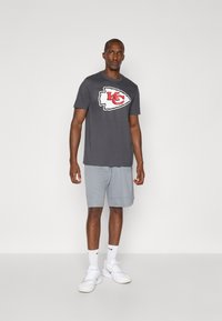 Grå T-shirt med et stort hvidt og rødt KC-logo, parret med lysegrå shorts. Model iført hvide sneakers og sokker.