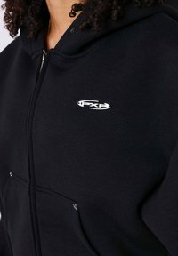 Sudadera negra con cremallera hecha de tela suave, con un logo en el pecho, un bolsillo delantero y un acabado texturizado.