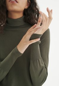 Pull à col roulé vert olive avec poignets côtelés et texture lisse, doté de manches longues ajustées. Détails des mains visibles.