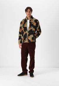 Giacca in pile con zip in un motivo camouflage con macchie verdi, marroni e nere; abbinata a pantaloni in velluto a coste marroni e sneakers nere.