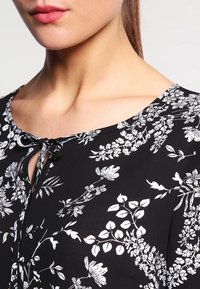 Blouse noire à motifs floraux avec des designs botaniques blancs, col en clé, et texture douce. Détails de tissu noué au col.