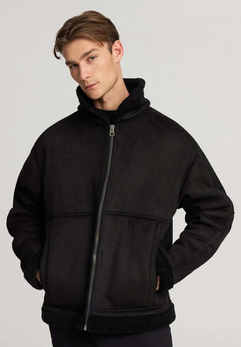 Giacca nera con zip e colletto alto, realizzata in materiale morbido e testurizzato. Presenta tasche frontali e dettagli in pelliccia a contrasto su polsini e orlo.