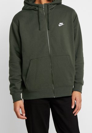 Mężczyzna ubrany w ciemnozieloną bluzę na zamek firmy Nike z przednimi kieszeniami oraz czarne spodnie, stojący na neutralnym tle.