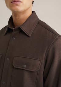 Barna gombos ing, gallérral, amely két mellzsebbel és fekete patentgombokkal rendelkezik. Az anyag sima, enyhén texturált felülettel.